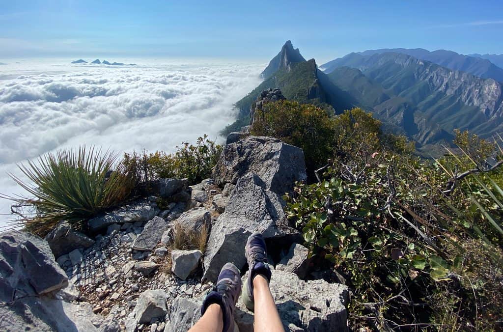 A hiker sits on top of La "M" summit above the clouds, with jagged mountain peaks in the distance.