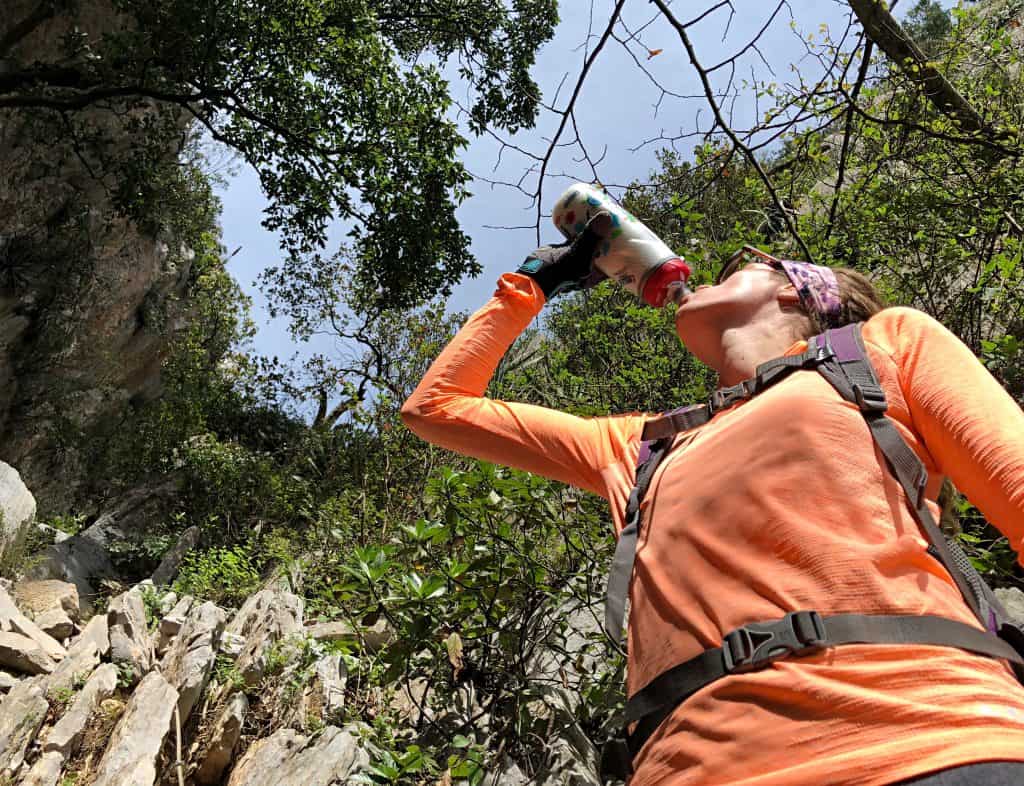 A hiker in a bright orange top drinking from a sports bottle with the rocky mountains of Chipinque in the background.