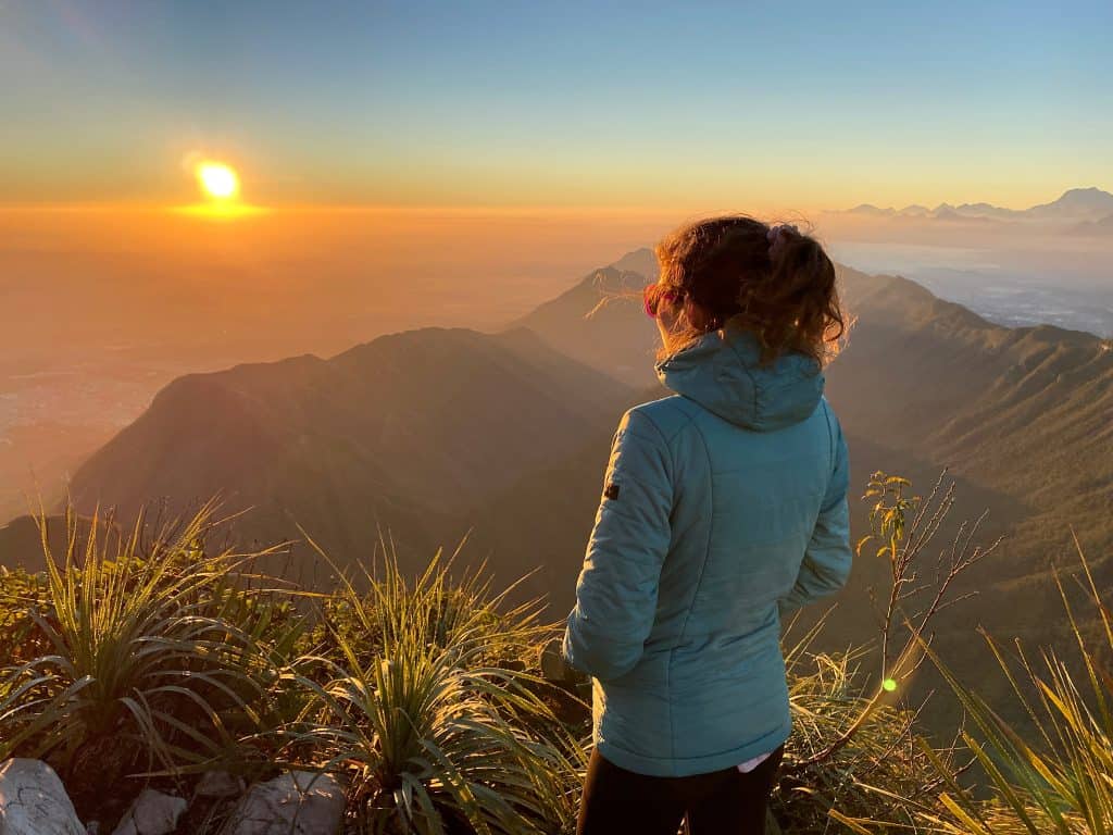 A hiker watching sunrise from the top of La Antena, Cerro de la Silla, Monterrey, with rolling mountains in the background.